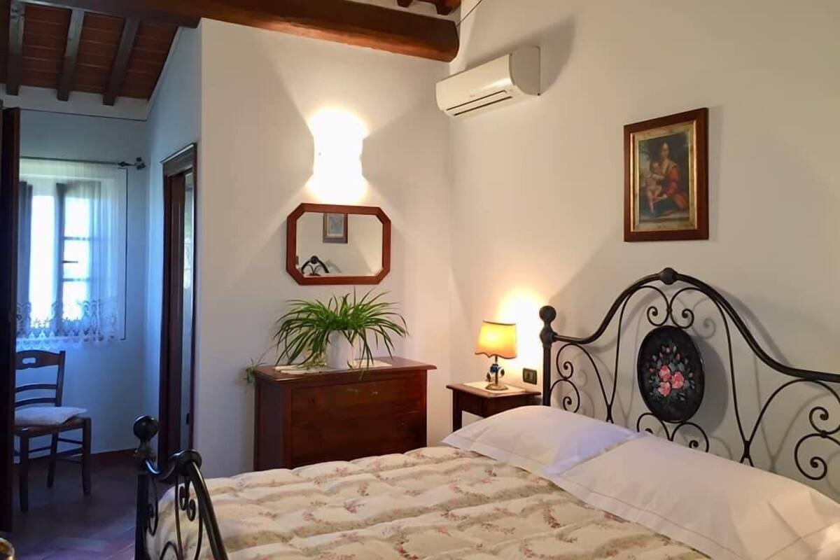 Un dormitorio con cama, cómoda, espejo, planta en maceta, lámpara y un cuadro en la pared.