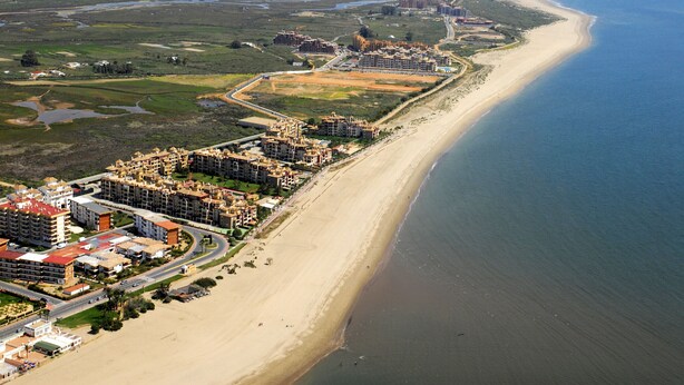 Luchtbeeld van een kustgebied met een lang zandstrand, een dicht cluster van meerverdiepingen en een jachthaven met boten.
