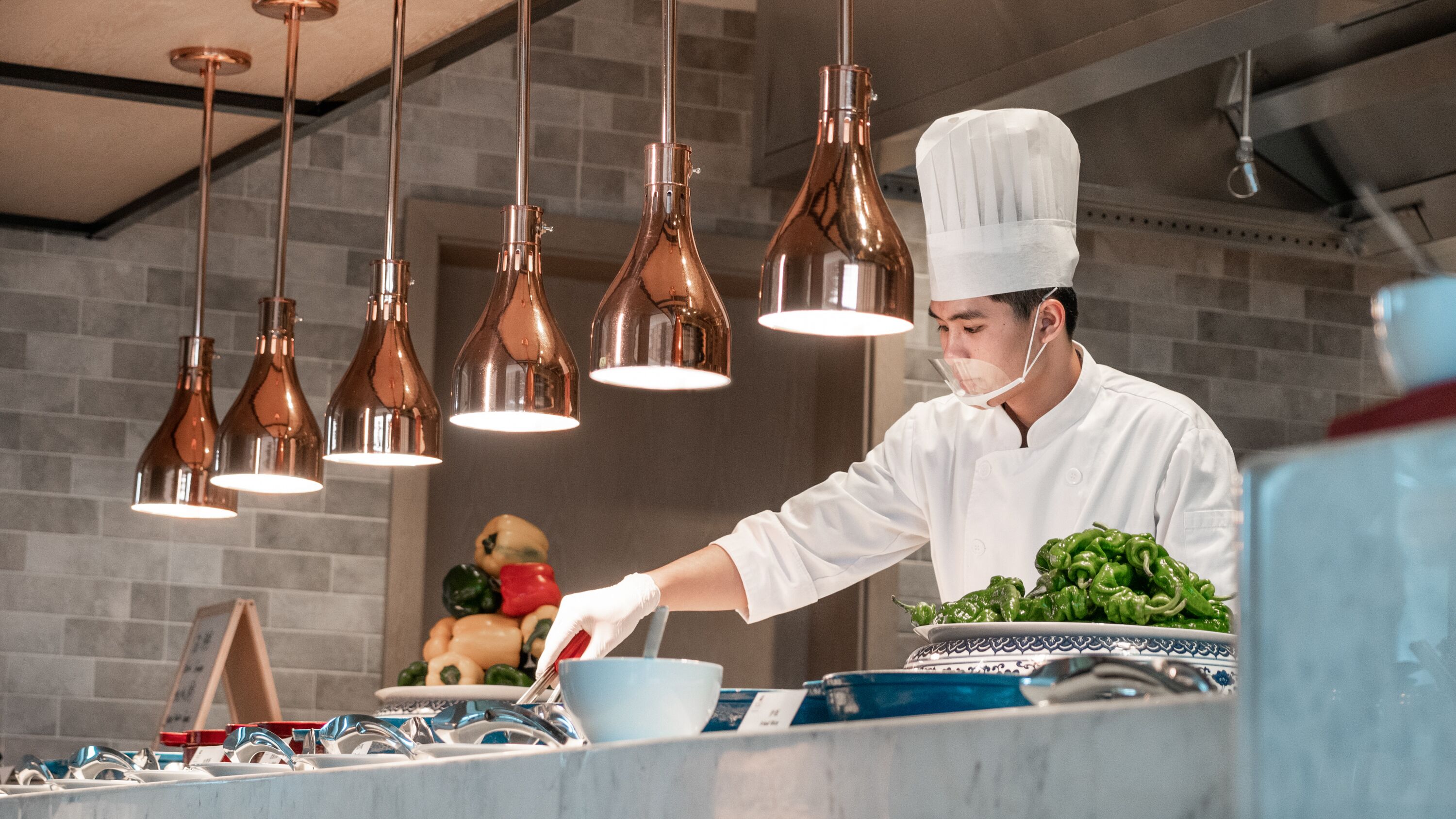 A chef in a professional kitchen with copper pendant lights, a tiled backsplash, and a variety of fresh vegetables on the counter.