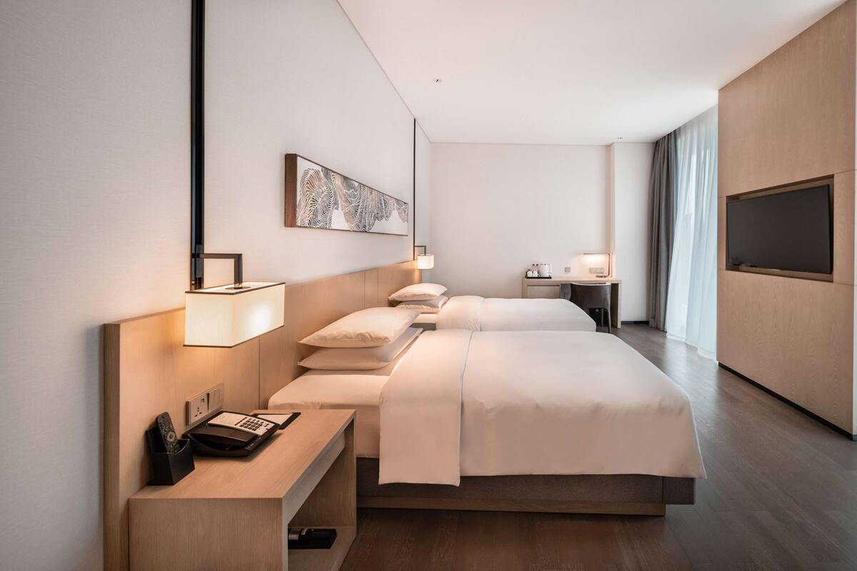 Quarto de hotel moderno com uma cama grande, mesas de cabeceira, escrivaninha, televisão e uma janela ampla com cortinas.