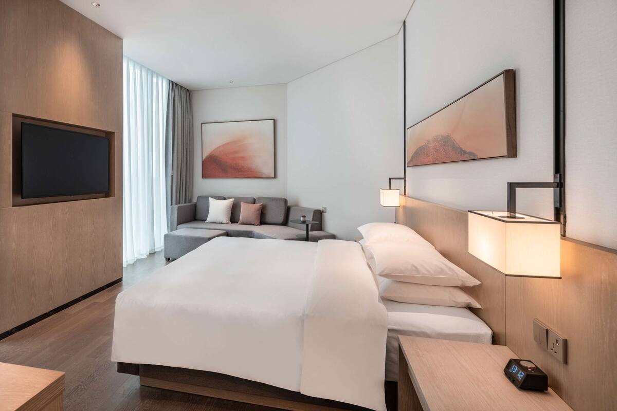 Quarto de hotel moderno com uma cama grande, um sofá, uma TV de tela plana e um quadro na parede.