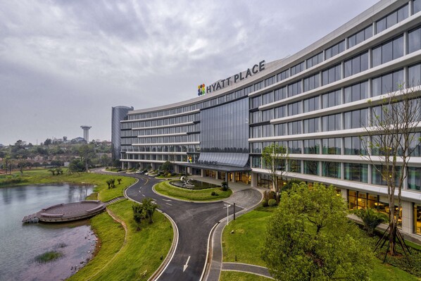 A modern hotel with a curved facade, a landscaped area with a water feature, and a view of the city skyline in the background. - Hyatt Place Changsha Airport (Changsha)