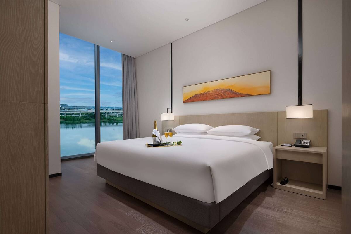 Quarto de hotel moderno com uma cama grande, mesa de cabeceira com abajur, quadro na parede e vista para um corpo de água através de grandes janelas.