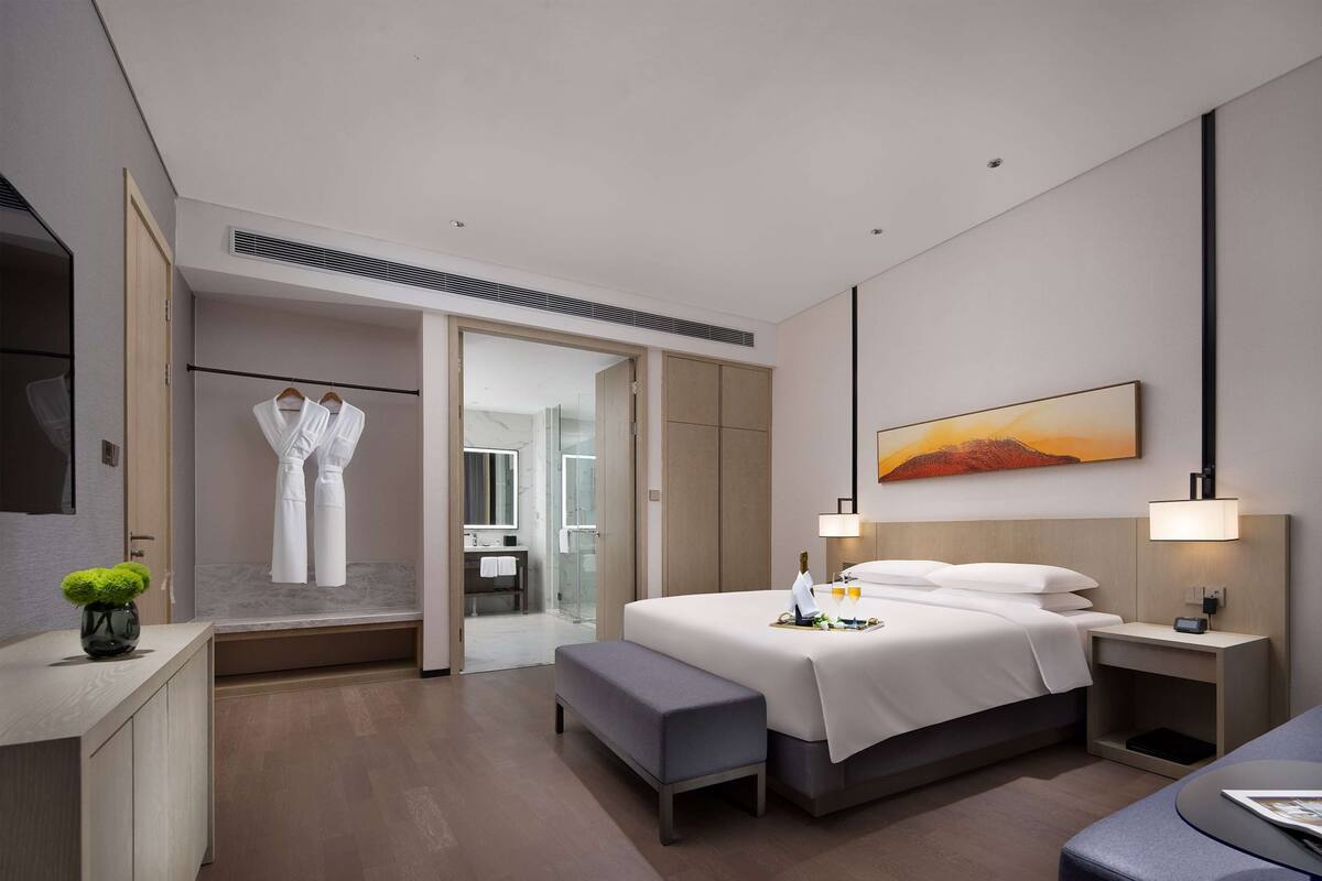 Quarto de hotel moderno com uma cama grande, mesas de cabeceira, um banco, uma escrivaninha e um banheiro visível através de uma porta aberta.