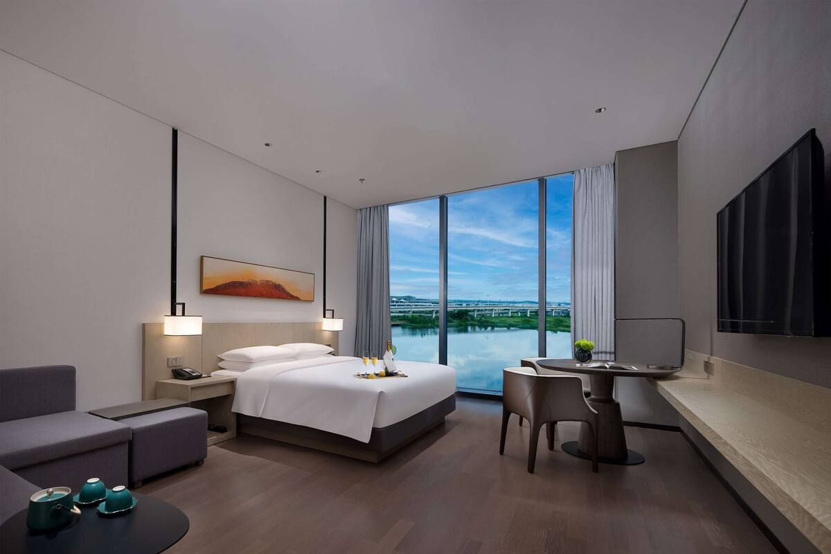 Quarto de hotel moderno com uma cama grande, uma escrivaninha, uma cadeira, uma televisão e vista para a cidade.
