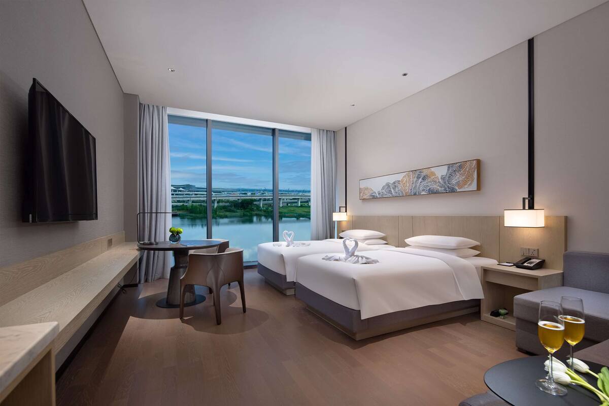 Quarto de hotel com duas camas, uma escrivaninha, uma cadeira, televisão e vista para um lago.