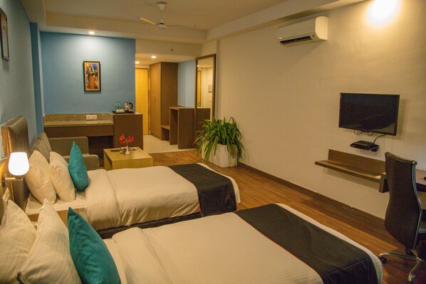 A hotel room with two beds, a desk, a chair, a TV, and a plant. - Four Leaf Service Suites (Roorkee)