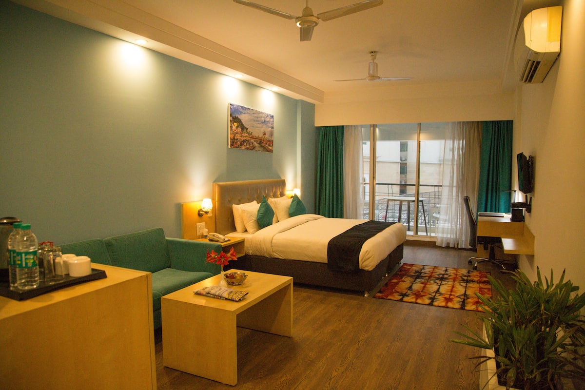 A hotel room with a bed, a sofa, a coffee table, a desk, a plant, and a view of the outside.