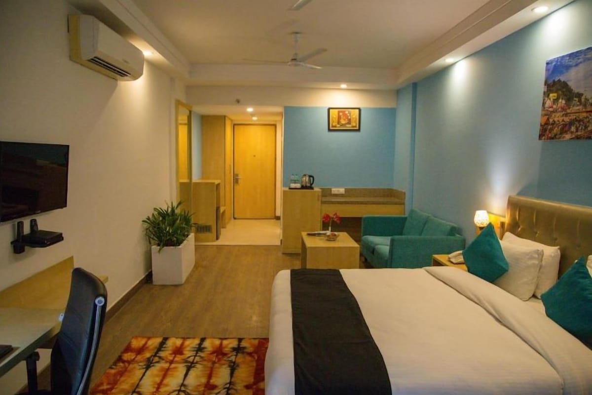 A hotel room with a bed, a desk, a chair, a TV, a plant, and a painting on the wall.