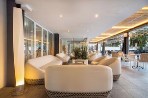 Un área de estar al aire libre con mobiliario moderno, sillas y mesas acolchadas, paredes de vidrio e iluminación lineal en el techo.