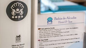 Dispensador de sabonete líquido com logotipo e texto indicando que é ecológico. Ao fundo, placa do 'Bahía de Alcudia Hotel & Spa' com logotipo e endereço do site.