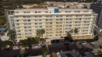 Un gran hotel con varios pisos y balcones, rodeado de coches aparcados y palmeras.