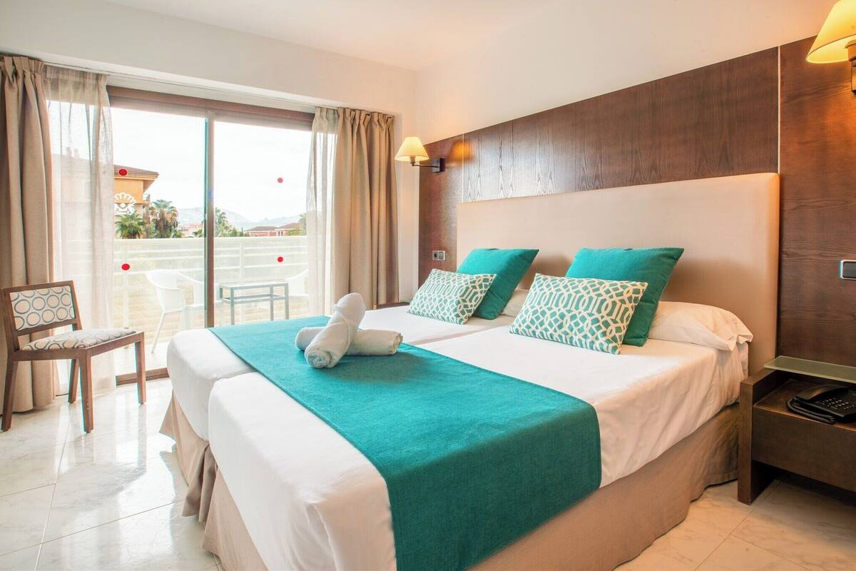 Een hotelkamer met een groot bed, een bureau met stoel, een balkon met uitzicht en een houten hoofdeinde.