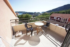 A balcony with a table and two chairs, a white suitcase, and a view of a cityscape with buildings and a hill in the background. - Apartments Becici Sunset (Becici)