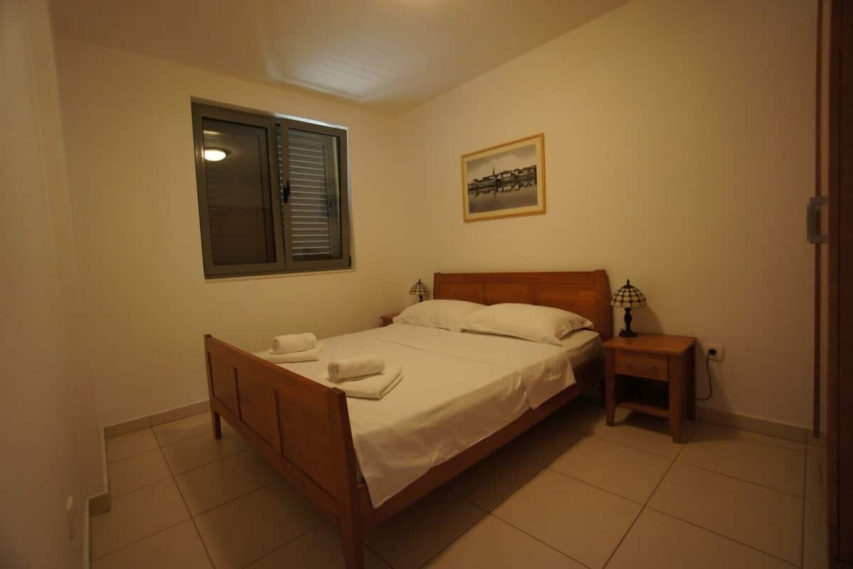 A bedroom with a wooden bed, white linens, a nightstand with a lamp, a window with blinds, and a framed picture on the wall.