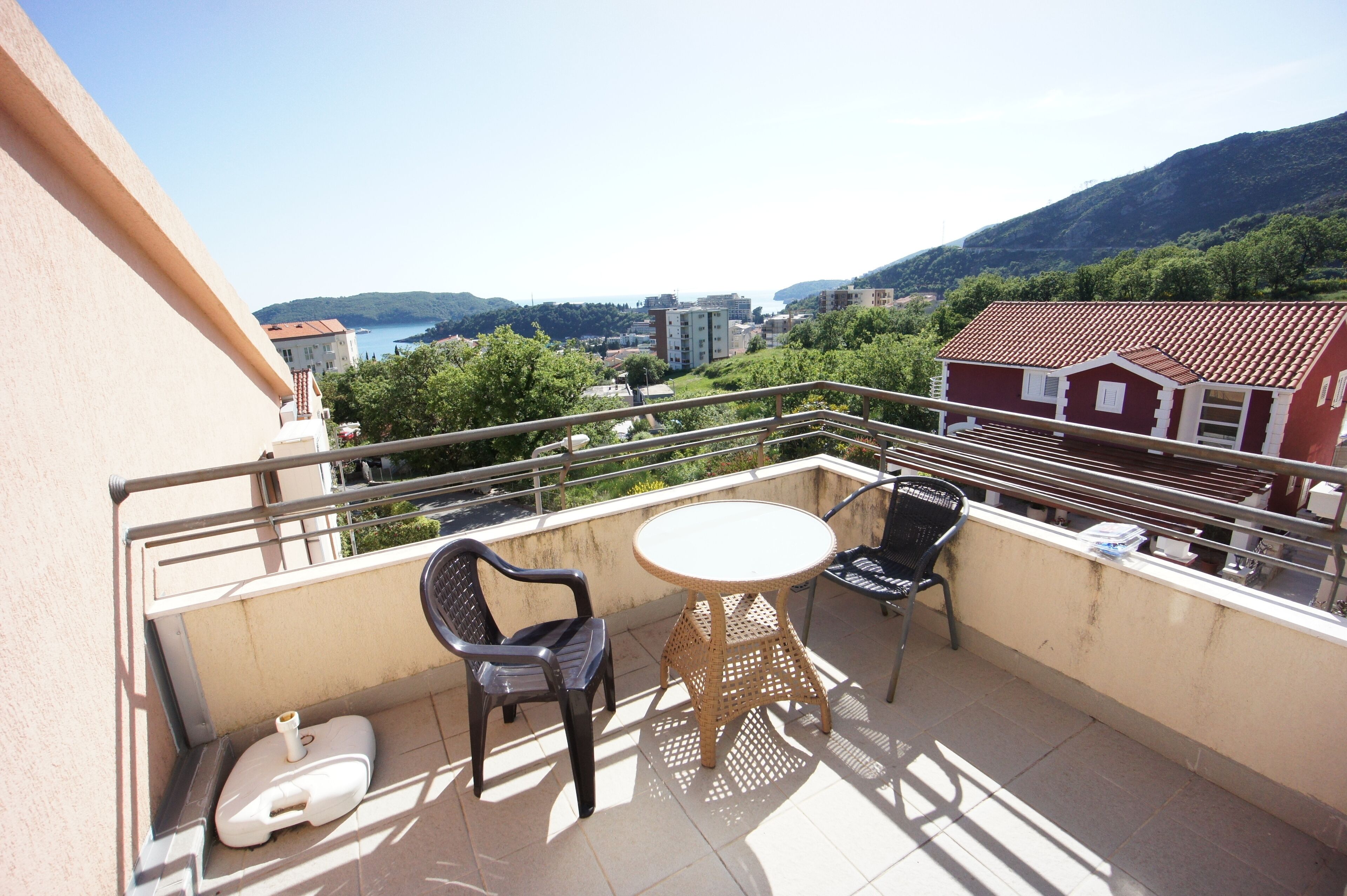 A balcony with a round table and two chairs, overlooking a scenic view of a coastal town and mountains.