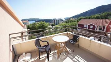 A balcony with a round table and two chairs, overlooking a scenic view of a coastal town and mountains.