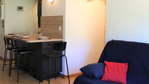 A compact living space with a dining area featuring a wooden table and chairs, a dark blue sofa with a red pillow, and a coat rack with hangers. - Ski-In/Ski-Out Apt, 2 Rooms, Mountain Corner, Sleeps 4, Central Vars, Near Slopes & Shops (Vars)