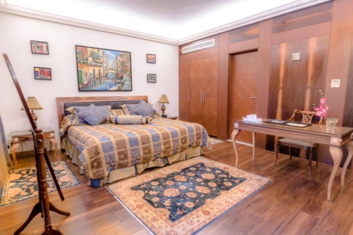 A bedroom with a bed, wooden wardrobe, desk, and a painting on the wall.