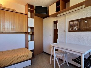 Quarto compacto com cama, mesa pequena, cadeira e televisão na parede.