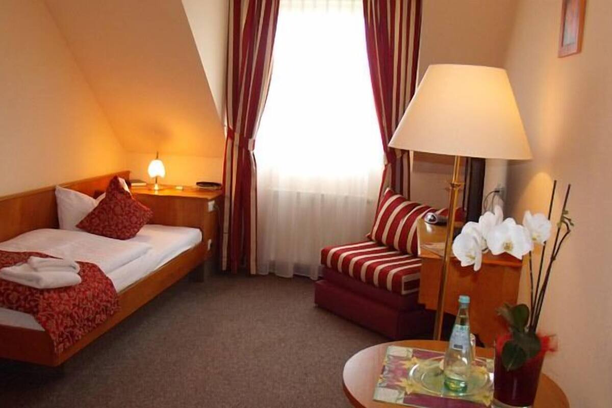 A hotel room with a bed, a chair, a desk with a lamp, and a small table with a bottle and flowers.