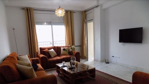 Ein Wohnzimmer mit einer Sofagarnitur, einem Couchtisch, einem gemusterten Teppich, einem an der Wand montierten Fernseher und einem Balkon mit Vorhängen.