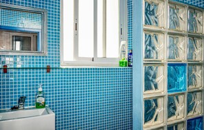 Ein Badezimmer mit blauen Mosaikfliesen, einem Fenster mit weißem Rahmen, einem Waschbecken mit Wasserhahn und einer Seifenflasche.