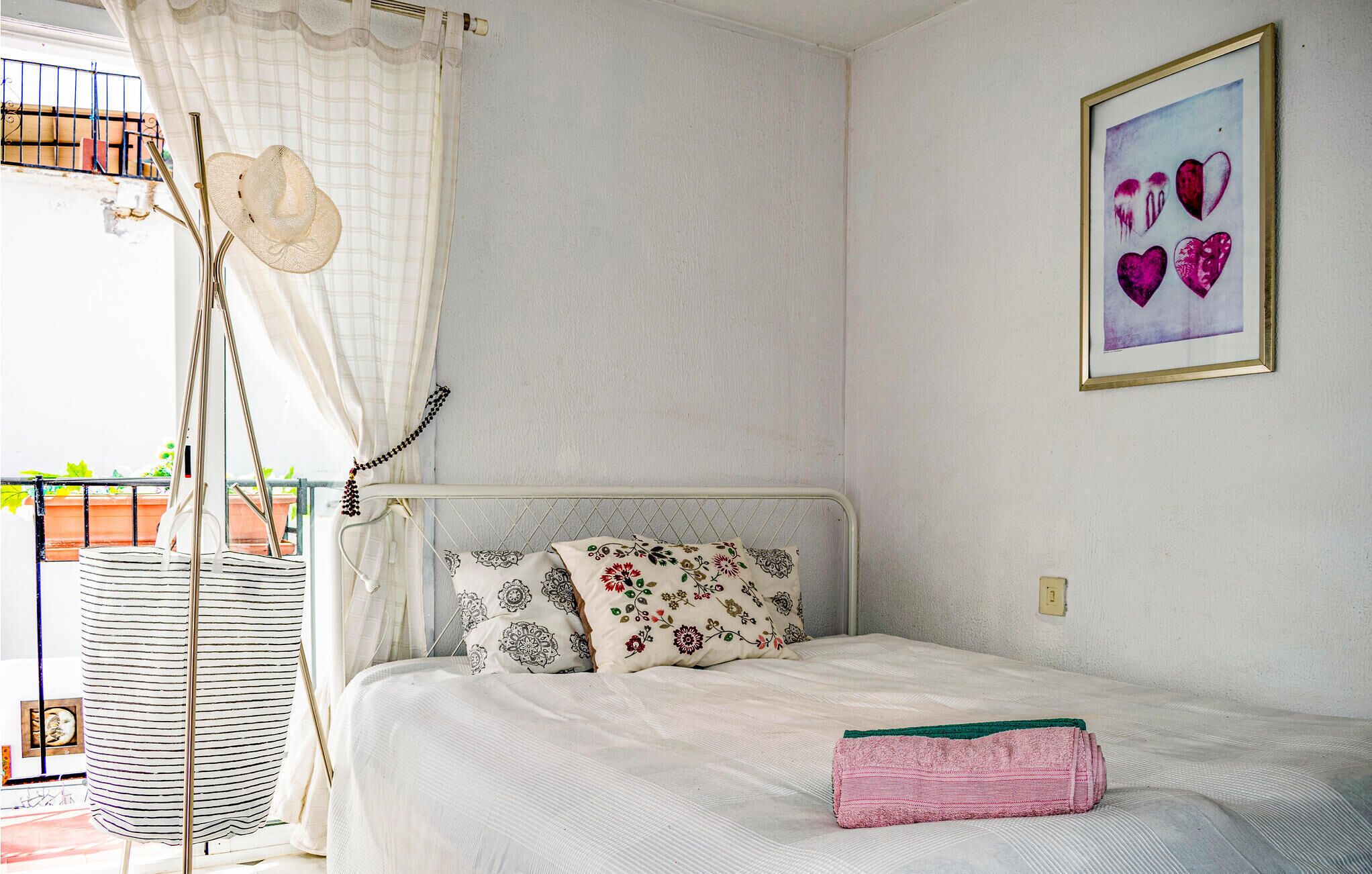 A bedroom with a bed, floral pillows, a pink towel, a framed artwork, and a balcony with a white curtain and a hat.