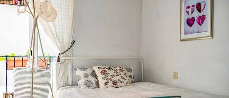 A bedroom with a bed, floral pillows, a pink towel, a framed artwork, and a balcony with a white curtain and a hat.