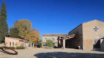 Un patio con edificios de piedra, un ciprés alto y una escultura de metal.