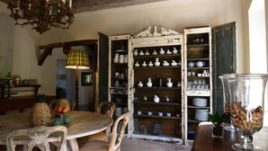 A rustic dining area with a wooden table, chairs, and a chandelier. There are shelves with dishes and a vase with corks. -  Commanderie de Peyrassol (Flassans-sur-Issole)