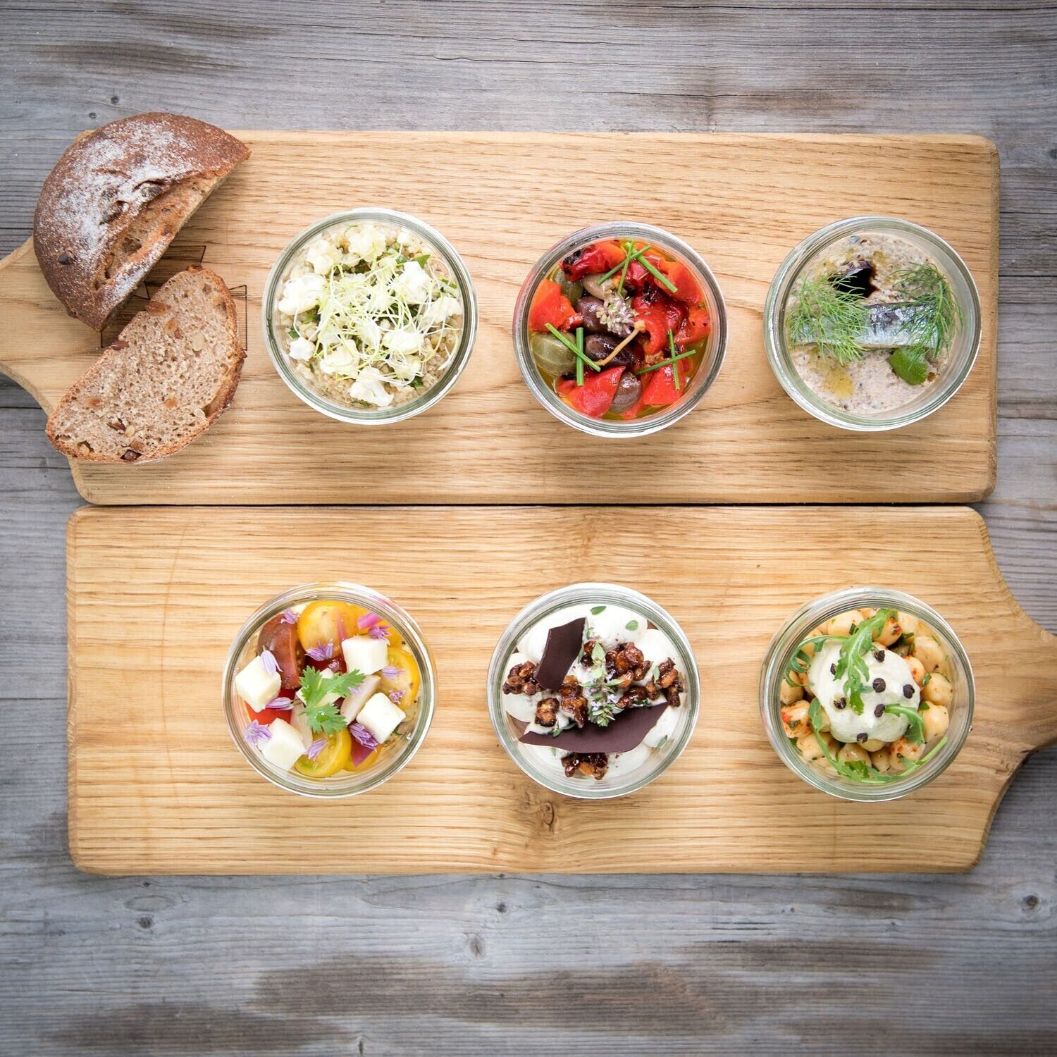 Six glass bowls of food on a wooden board, with slices of bread on the side.