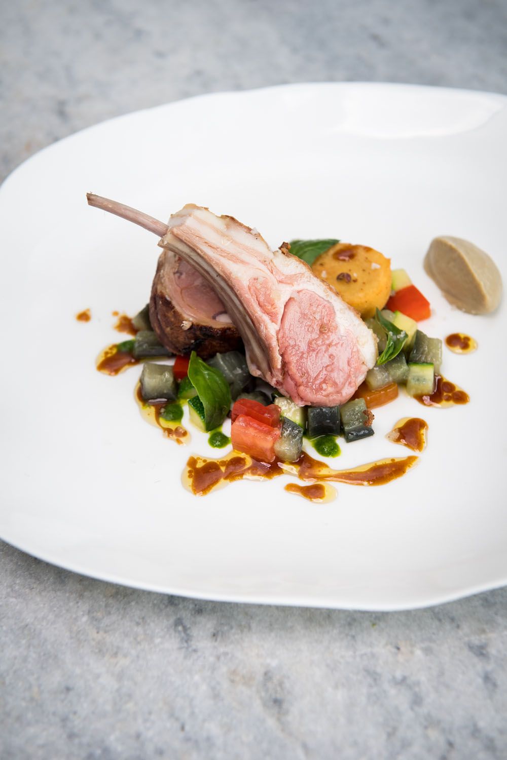 Une assiette blanche contenant une côte d’agneau, des légumes et une sauce.