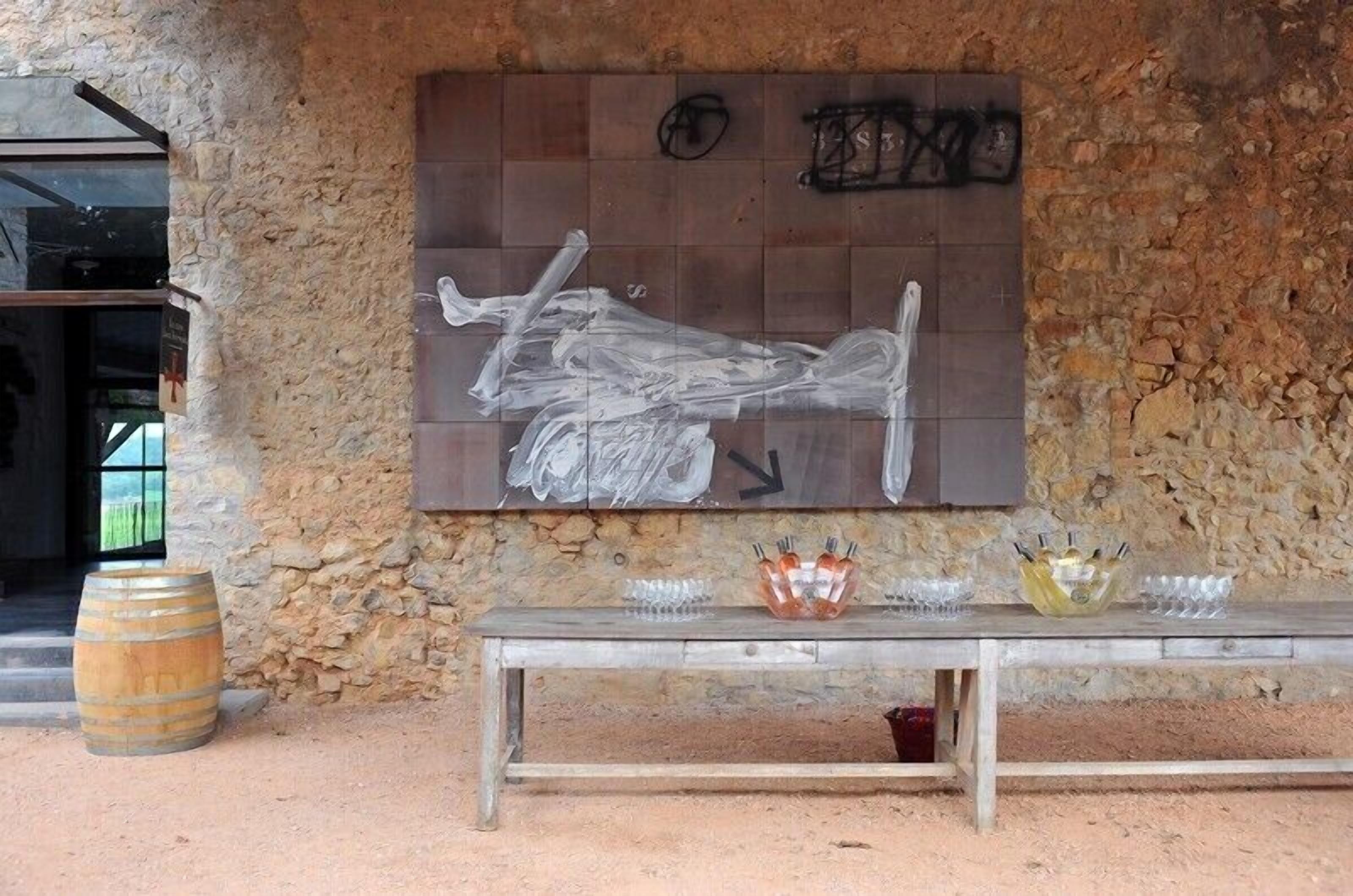 A rustic stone wall with a large abstract painting, a wooden bench with glassware, and a barrel.