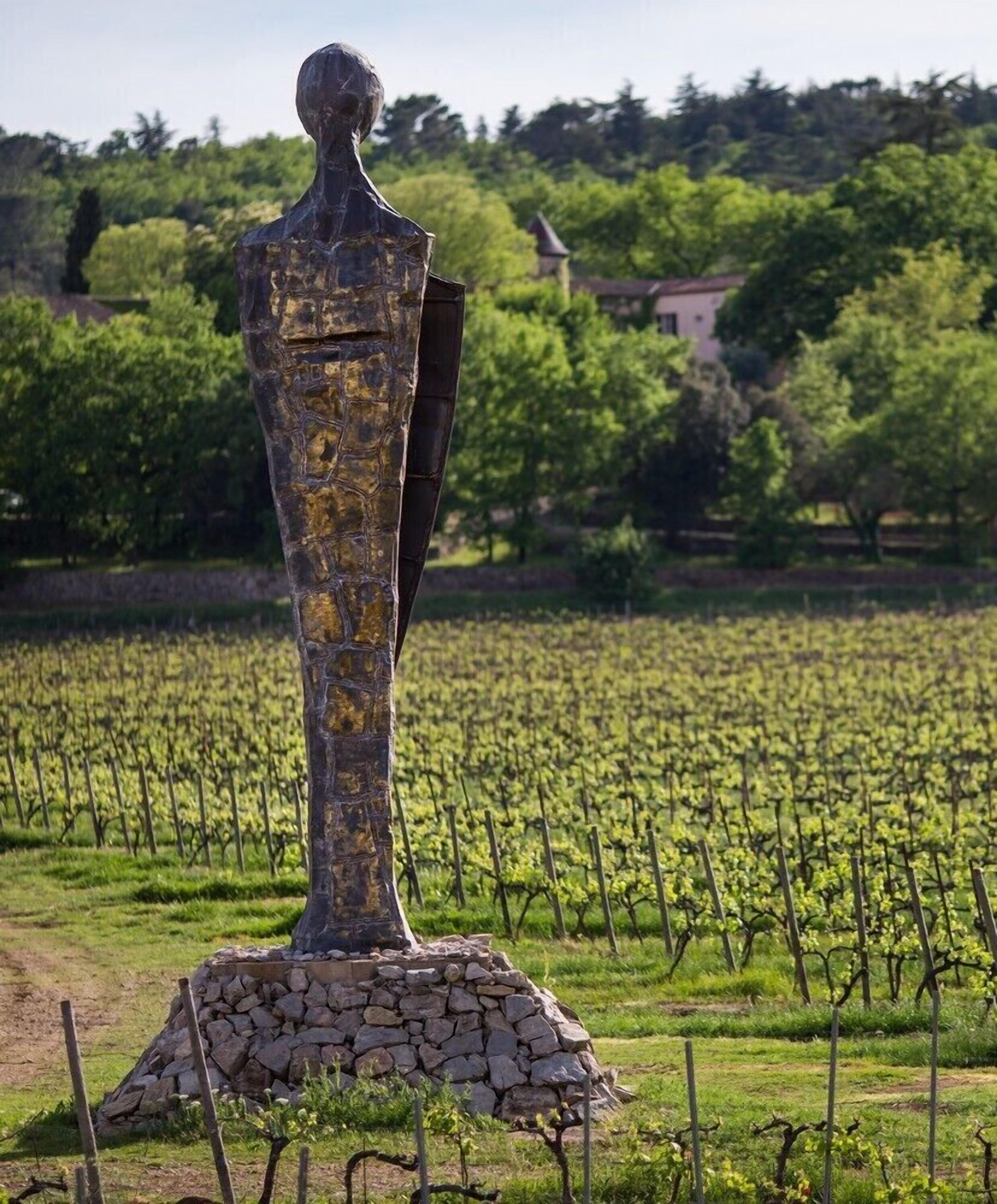 A sculpture in a vineyard with a stone base, surrounded by rows of grapevines and trees in the background.