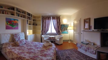 Ein Schlafzimmer mit Bett, Schreibtisch, Stuhl, Bücherregal, Fernseher und einem Fenster mit Vorhängen.