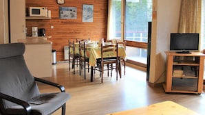 Un salon chaleureux aux murs en bois, une table à manger dressée pour quatre personnes, un canapé, un téléviseur posé sur un meuble et une fenêtre avec des rideaux.