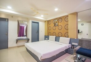 A hotel room with a bed, a sofa, a small table with a remote control, a mirror, and a ceiling fan. - Hotel Morya Regency (Bhopal)