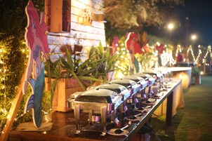 Una hilera de estaciones de bufé con recipientes de comida y utensilios, frente a un fondo de luces colgantes y adornos.