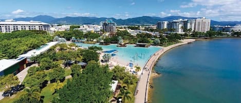 An aerial view of a coastal resort with a large pool area, beachfront promenade, and surrounding greenery.