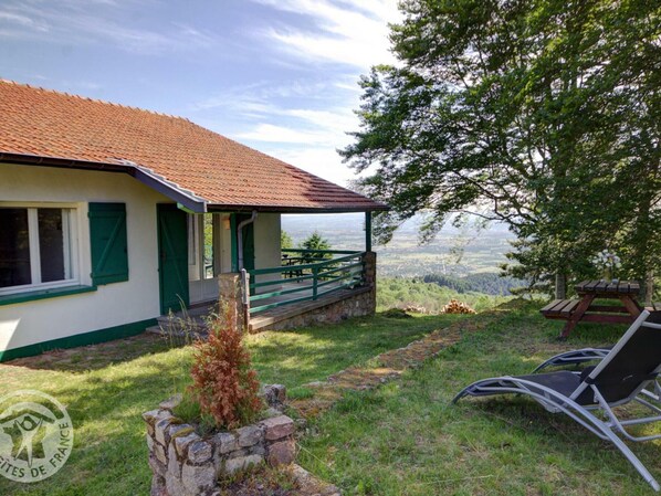 A chalet with a stone foundation, green shutters, and a red-tiled roof. It features a stone bench and a picnic table, surrounded by lush greenery and a view of the landscape.