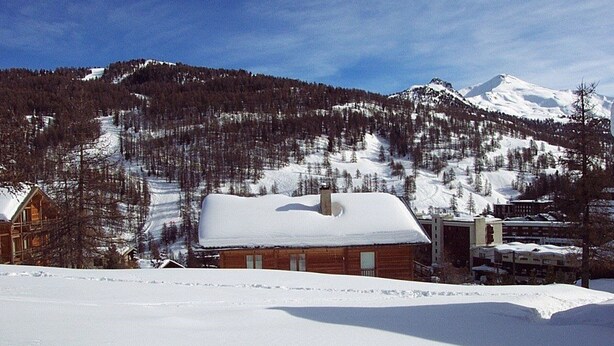Une cabane en bois dont le toit est recouvert de neige, entourée d’un paysage enneigé et avec une montagne en arrière-plan.