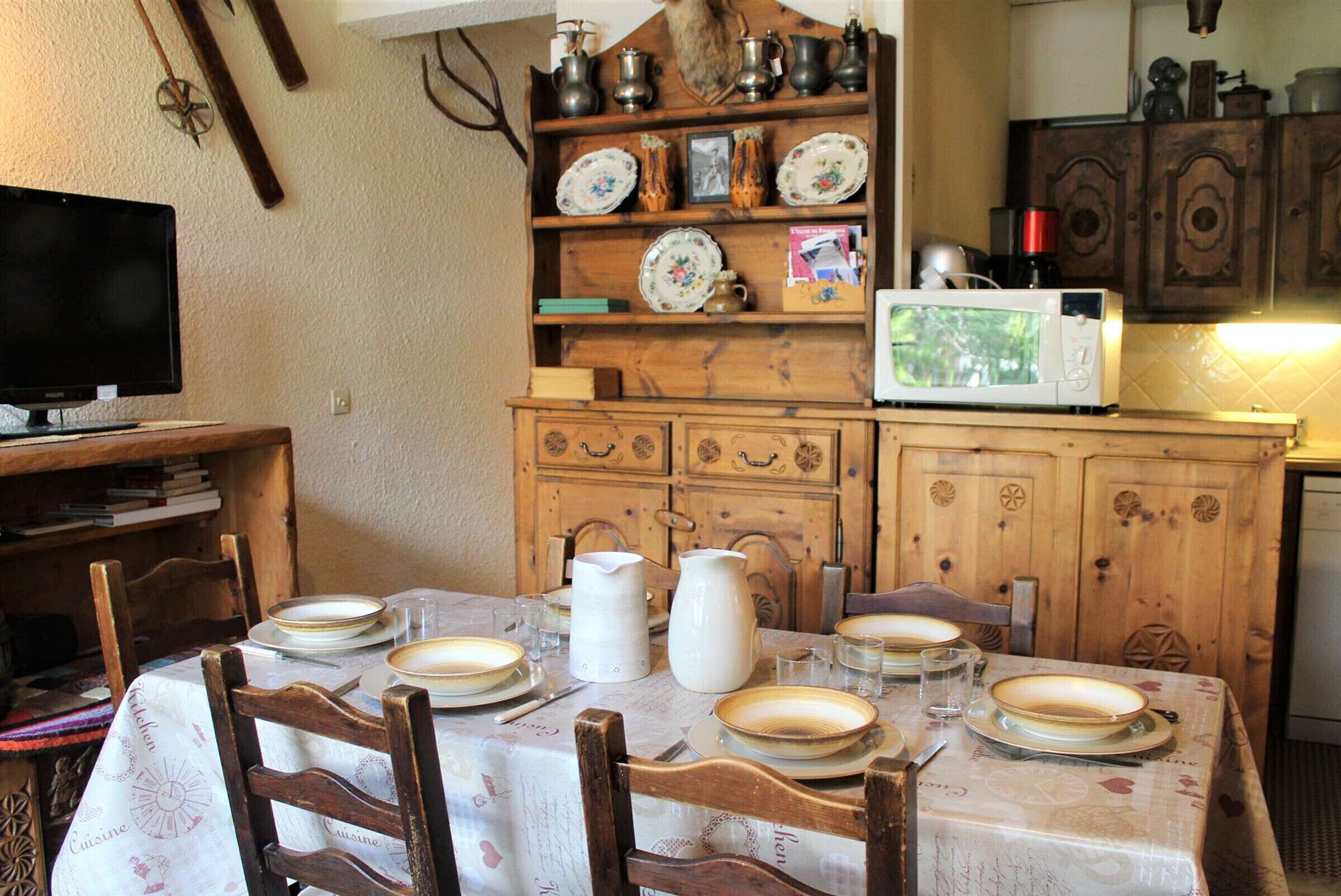 A dining area with a wooden table set for four, surrounded by chairs. A television is mounted on the wall to the left.