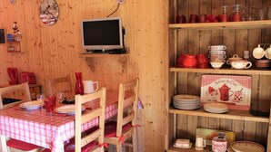 Une salle à manger en bois avec une table dressée pour quatre personnes, un téléviseur posé sur une étagère et une collection d’ustensiles de cuisine rouges et blancs disposés sur des étagères.
