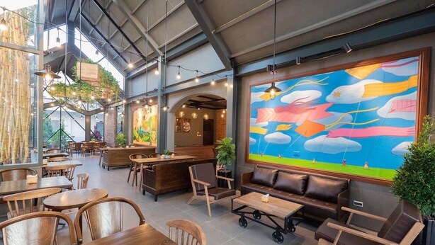 Un café spacieux avec des tables et des chaises en bois, une grande fresque murale et des plantes suspendues.
