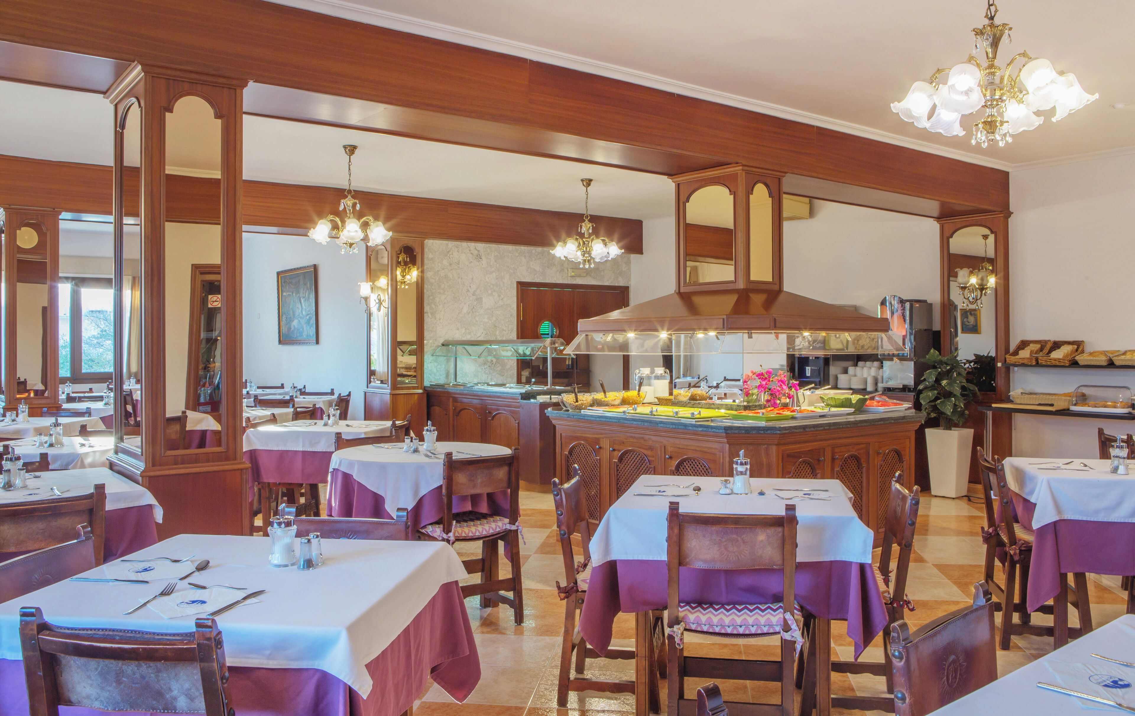 A restaurant with multiple dining tables, a central buffet, and chandeliers.