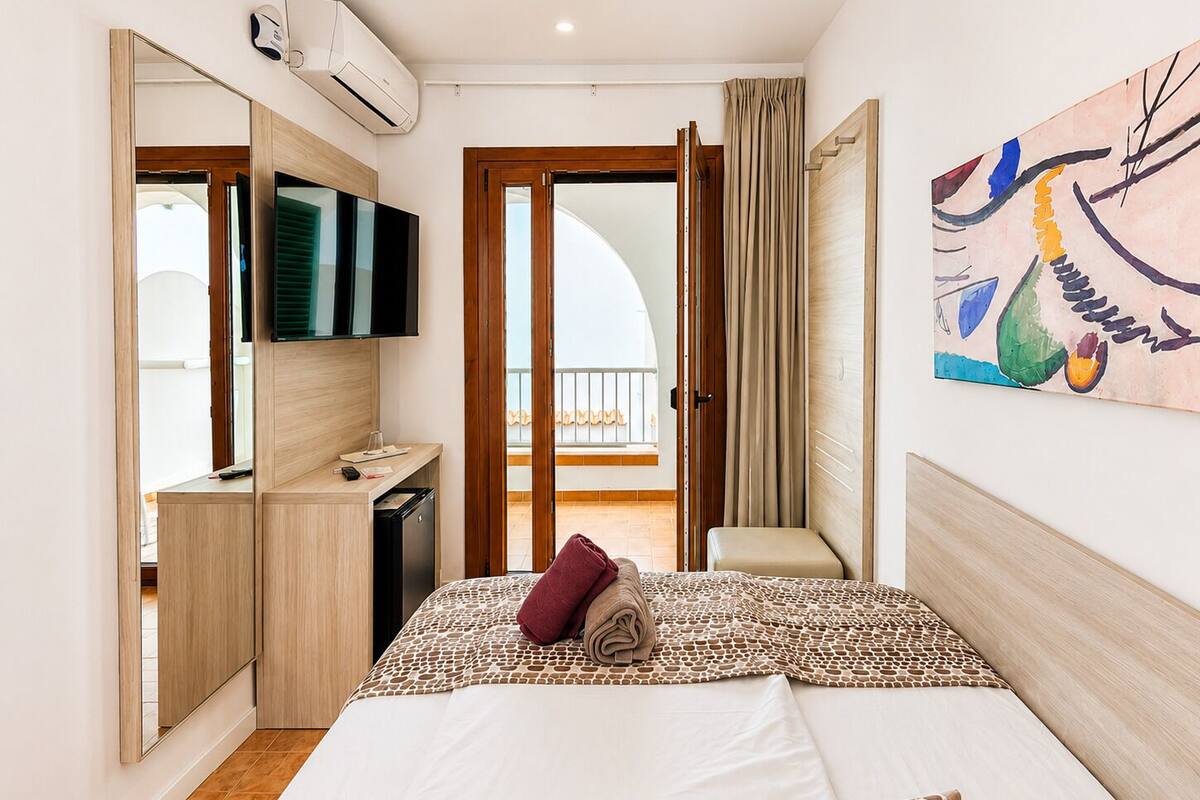 A hotel room with a bed, a TV, a desk, a wardrobe, a balcony, and a painting.