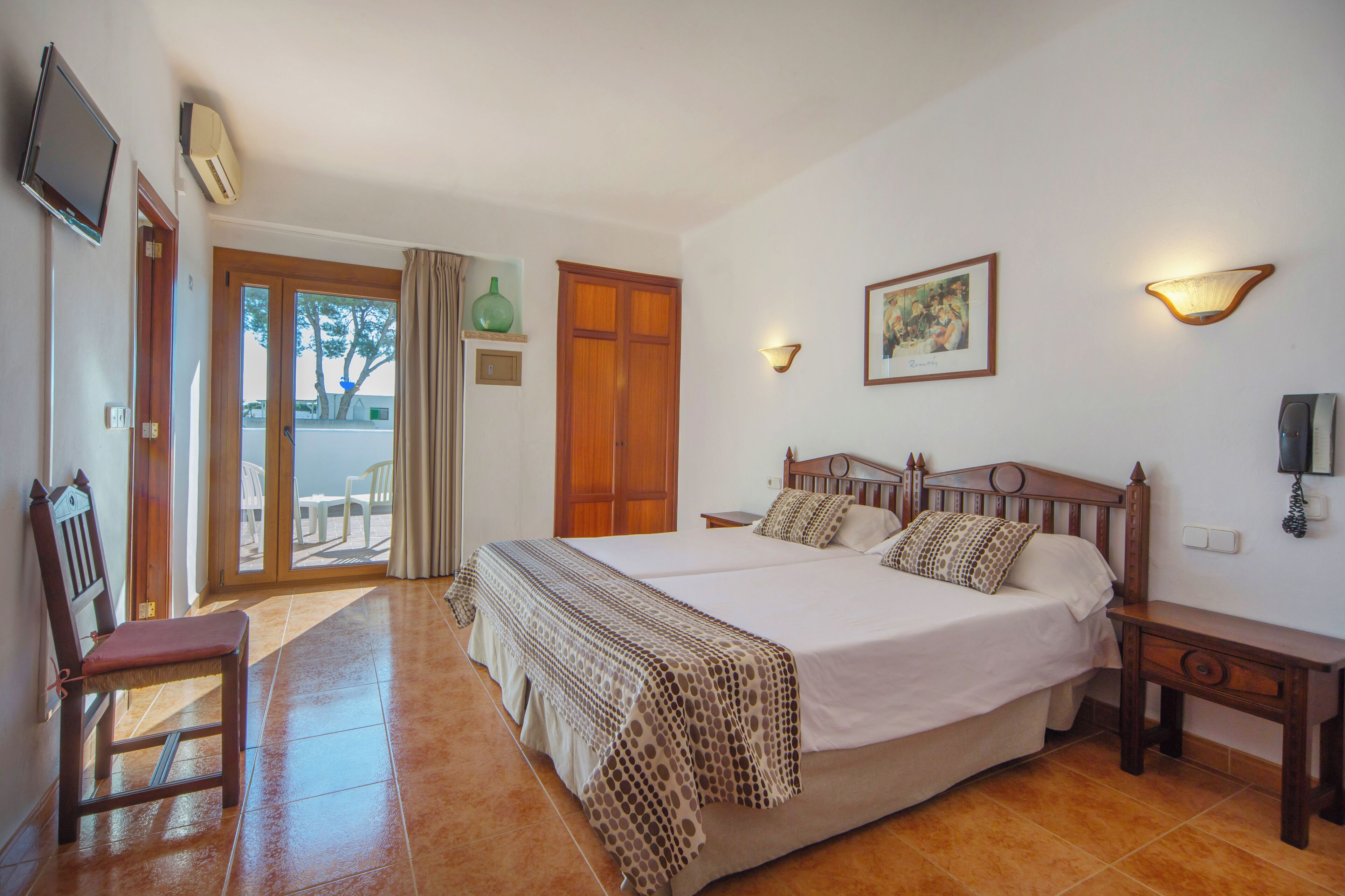 Uma cama de casal com cabeceira de madeira, mesa de cabeceira e uma cadeira. Uma porta dá acesso a uma área externa com vista para árvores.
