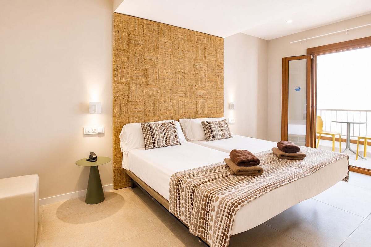 A double bed with a patterned bedspread, a bedside table with a lamp, and a view of a balcony with outdoor furniture.