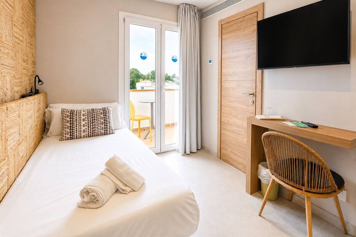 A hotel room with a bed, a desk, a chair, a TV, and a sliding glass door to a balcony.